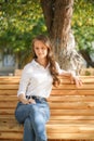 A girl in a white blouse and blue jeans sits on a wooden bench in autumn Park square Royalty Free Stock Photo