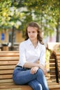 A girl in a white blouse and blue jeans sits on a wooden bench in autumn Park square Royalty Free Stock Photo