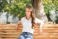 A girl in a white blouse and blue jeans sits on a wooden bench in autumn Park square Royalty Free Stock Photo