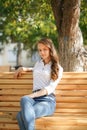 A girl in a white blouse and blue jeans sits on a wooden bench in autumn Park square Royalty Free Stock Photo