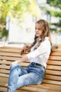 A girl in a white blouse and blue jeans sits on a wooden bench in autumn Park square Royalty Free Stock Photo