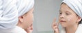 Girl in a white coat with a towel on her head moisturizes face with a cream in front of a mirror