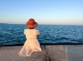 a girl in a white dress and a hat sits on the pier and looks at the sunset sea, rear view Royalty Free Stock Photo