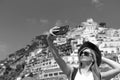 A girl in a white dress makes selfie photo on smartphone on the beach in Positano. View of houses and hotels in the background. Royalty Free Stock Photo