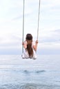 Girl in white dress is riding swing above water. Back view. Blue sky horizon and sea Royalty Free Stock Photo