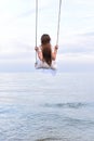 Girl in white dress is riding swing above water. Vertical frame. Back view. Background of blue sky horizon and sea Royalty Free Stock Photo