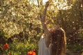 Girl in white dress sitting among flowers and fluff with threw back her head near tulips in sunset, dandelions and cherry flowers