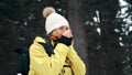 A girl in a white hat and a yellow anorak warms her hands in the winter in the forest