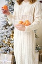 A girl in a white knitted dress stands against the backdrop of a Christmas tree and holds a basket of tangerines in her Royalty Free Stock Photo