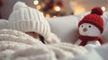 A girl in a white knitted hat covered with a blanket sits on the sofa next to a toy snowman, Christmas concept Royalty Free Stock Photo