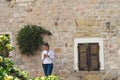 Girl with white long shirt sending a message in smart phone and stone house with flowers in the facade. Woman typing in cell phone