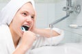 A girl in a white robe and a towel on her head, sitting in the bathroom and brushing her teeth over the sink
