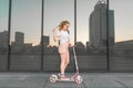 Girl in a white shirt with a tail and standing on a pink scooter on a background of dark mirrored wall Royalty Free Stock Photo