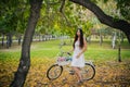 Girl in a white skirt and a vest with a bicycle and a basket of flowers stands on yellow leaves in the park in the evening, in the Royalty Free Stock Photo