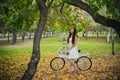 Girl in a white skirt and a vest with a bicycle and a basket of flowers stands on yellow leaves in the park in the evening, in the Royalty Free Stock Photo