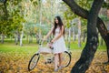 Girl in a white skirt and a vest with a bicycle and a basket of flowers stands on yellow leaves in the park in the evening, in the Royalty Free Stock Photo