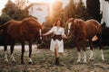 A girl in a white sundress and hat stands next to horses in nature in the eagle stables