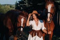 A girl in a white sundress and hat stands next to horses in nature in the eagle stables