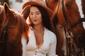 A girl in a white sundress and hat stands next to horses in nature in the eagle stables