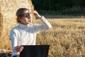 Girl in a white sweater against the background of a haystack with a laptop in her hands sits on the ground, looks into the distanc Royalty Free Stock Photo