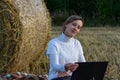 Girl in a white sweater on the background of a haystack with a laptop in her hands sits on the ground, looks at the camera Royalty Free Stock Photo