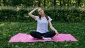 A girl in a white T-shirt and black leggings on a pink mat sits on a summer green lawn and performs a neck stretch