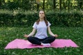 A girl in a white t-shirt and black leggings on a pink mat sits in a yoga pose on a summer green lawn