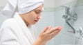 A girl with a white towel on her head and in a dressing gown, in the bathroom, washes the foam, for cleansing, from the face