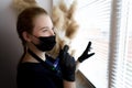 Girl who is a master of permanent makeup in a black mask and gloves holds a tattoo machine in her hands stands at the window and Royalty Free Stock Photo