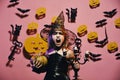 Girl with wild face on pink background with spooky decor. Kid in spooky witches costume holds carved pumpkin and Royalty Free Stock Photo