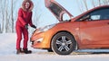 A girl in a red winter red suit stands next to a broken car.