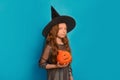 Cute kid in witch costume holding Halloween pumpkin or Jack o lantern pumpkin and looking at camera on blue background. Royalty Free Stock Photo