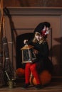 A girl in a witch costume sits near the fireplace on a pumpkin, holding a lantern in her hands Royalty Free Stock Photo