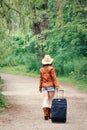 girl woman in leather jacket, blue denim shorts, straw hat, standing walking on country road wild forest with travel bag Royalty Free Stock Photo