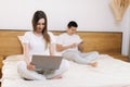 Girl working on laptop sitting in bed. Guy uses phone behind his girlfriend. Royalty Free Stock Photo