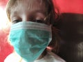 Girl 6 years old in a protective surgical mask on a red-brown background, lit by sunlight. The fight against the virus. Sad gray