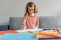 Girl of 8 years sitting on sofa at home drawing writing with pencil in notebook. Child blonde with glasses studying at home