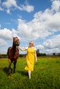 Girl in yellow dress with a horse on a green field and a blue sky with white clouds on the background Royalty Free Stock Photo