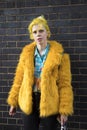 A girl with yellow hair in a faux fur coat, blue shirt with palm trees and skinny jeans poses against a black brick wall. Street Royalty Free Stock Photo