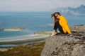 Girl in yellow jacket stitting on stone in on the edge of rocks in mountains and looking to camera