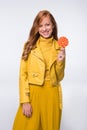 smiling redhead stylish girl in yellow leather jacket holdig lollipop