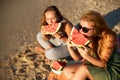 Girlfriends laughing and eating fresh watermelon at seaside in the morning. Two happy women friends sitting on towel and Royalty Free Stock Photo