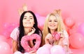 Girls lay near balloons, holds heart toys, pink background. Sisters, friends in pajamas at pajamas party. Blonde and