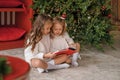 Girls in nightgowns is sitting on the floor by a Christmas tree in the early December morning and reading a book