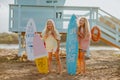 Girls posing with surfboards against blue lifeguard tower on the sand beach.
