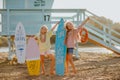 Girls posing with surfboards against blue lifeguard tower on the sand beach.