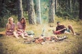 Girls sitting next to campfire while reading books. Bearded guy lying on grass in forest. Friends camping in summertime