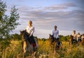 Girls in white shirts are riding horses on a blooming meadow in the rays of the setting sun.