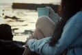 A girlsitting on sea rock, holding a cup of morning tea or coffee in the beach. Self care, relax weekend concept.