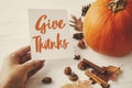 Give thanks text on blank card in hand on background of pumpkin, autumn leaves, anise, acorns on white wood. Happy thanksgiving
